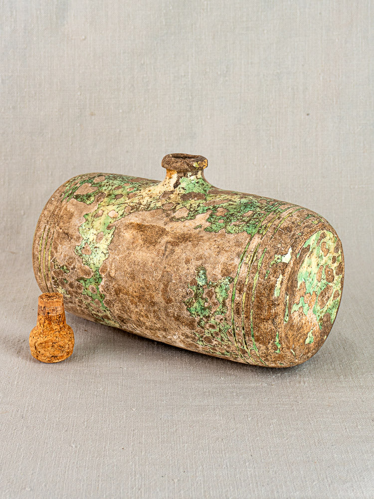 18th-century French terracotta bouillotte with weathered brown and green glaze, stopper alongside on neutral fabric background.