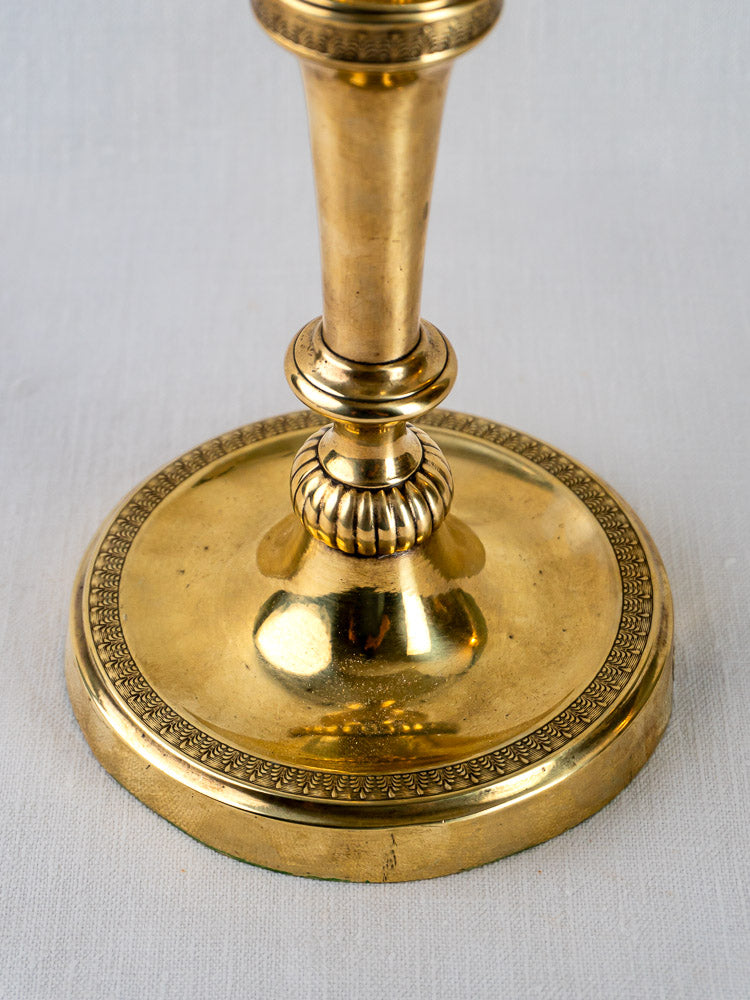 Ornate base of Empire Palmette brass candlestick with decorative edging on white textured surface.