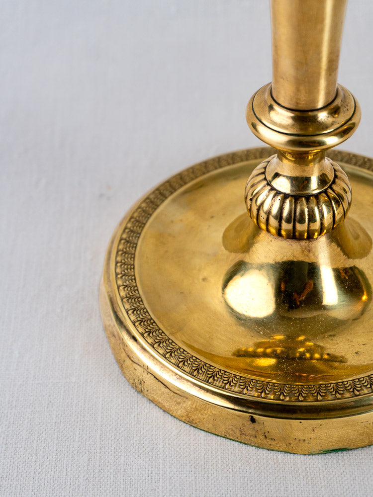 Ornate gold-tone base of Empire Palmette brass candlestick with beaded edge on white fabric.