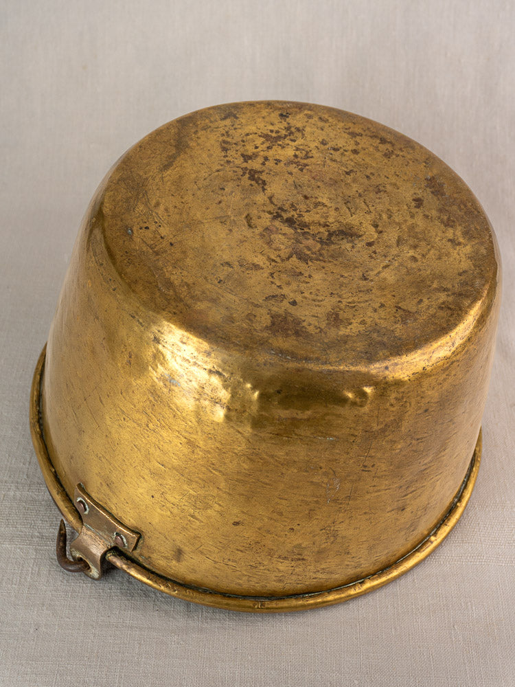 Antique French brass jam pan upside down on light cloth, highlighting worn round surface and metal clasp.