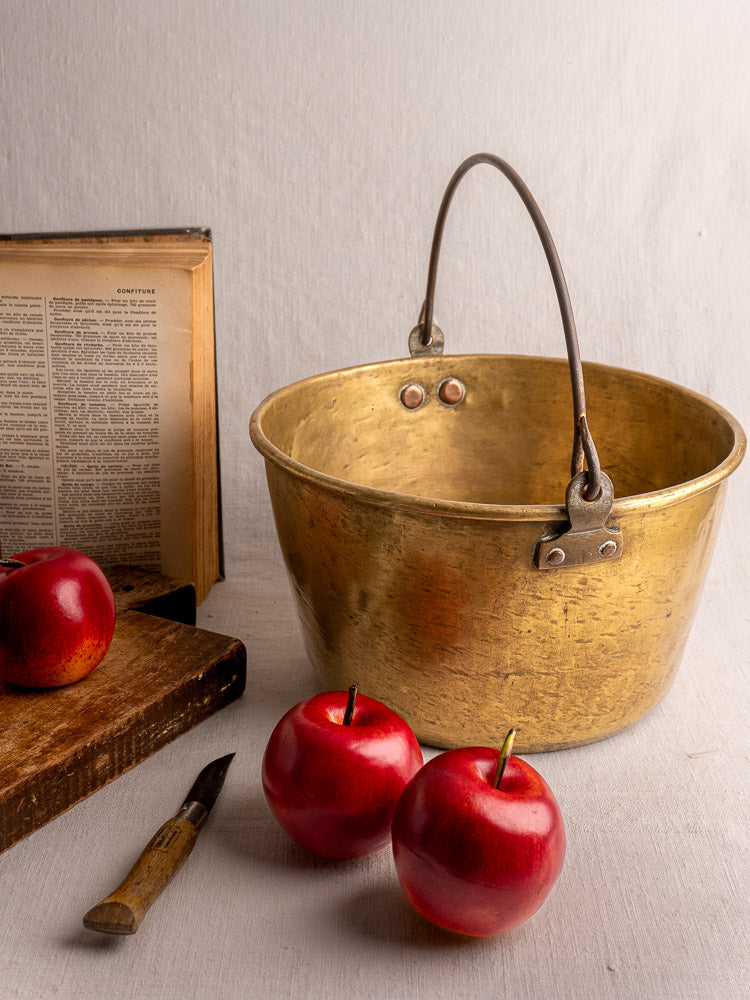 Antique French brass jam pan on a white surface with red apples, open book, cutting board, and small knife nearby.