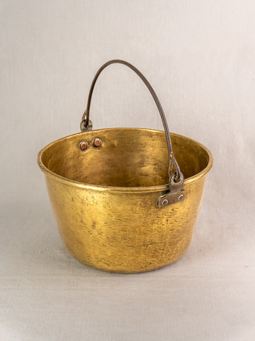 Antique French brass jam pan with rounded shape, dark metal handle, textured patina and rivets on beige background.