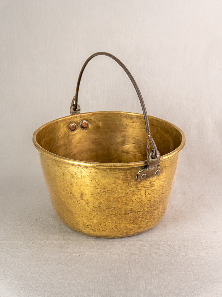 Antique French brass jam pan with rounded shape, dark metal handle, textured patina and rivets on beige background.