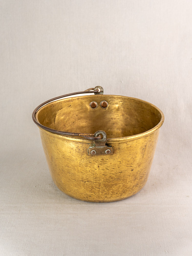 Antique French brass jam pan with metal handle and rivets, aged patina, on neutral fabric background.