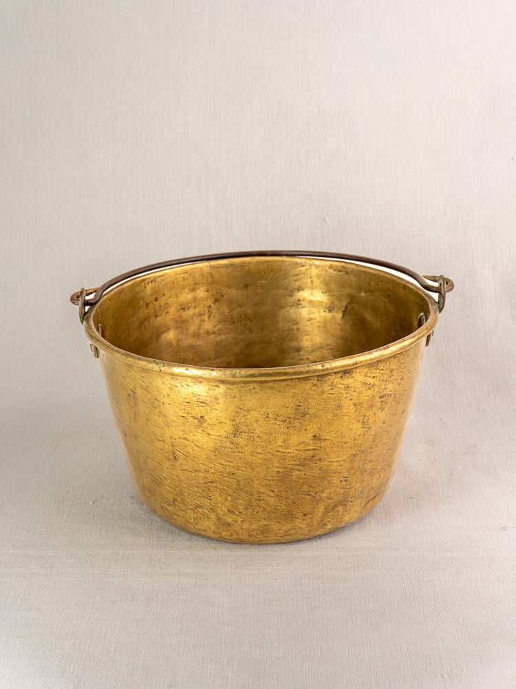 Antique French brass jam pan with metal handle, round shape, and gently worn patina on light fabric, beige background.