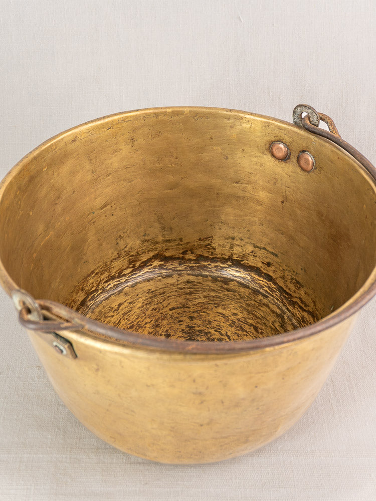 Antique French brass jam pan with textured, worn interior and two handles on a pale fabric surface.