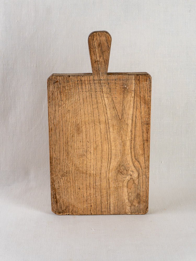 Antique French chestnut cutting board, circa 1900, with handle and rustic wood grain on pale fabric.
