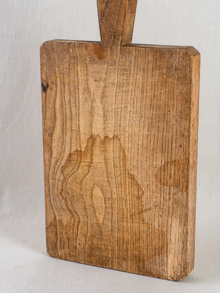 Antique French chestnut cutting board c. 1900 with handle, upright against light fabric background.