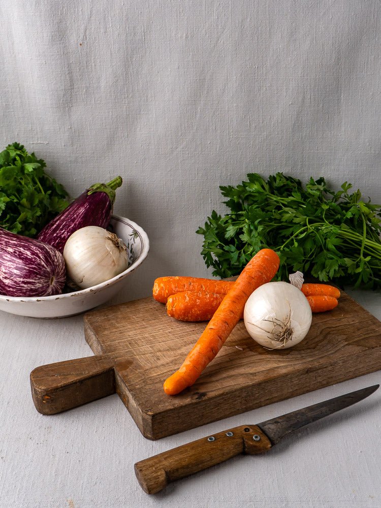 Antique French chestnut cutting board with carrots and onion, knife, and fresh parsley on fabric.