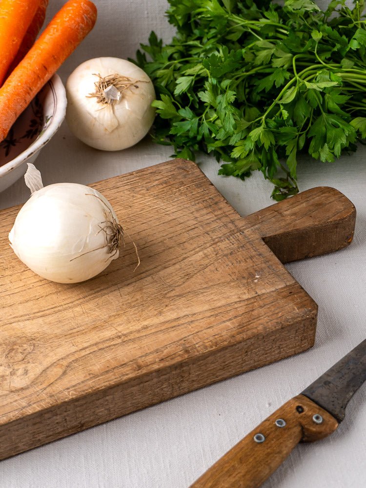 Antique French chestnut cutting board c. 1900 with white onion and knife; carrots, onions, and parsley behind.