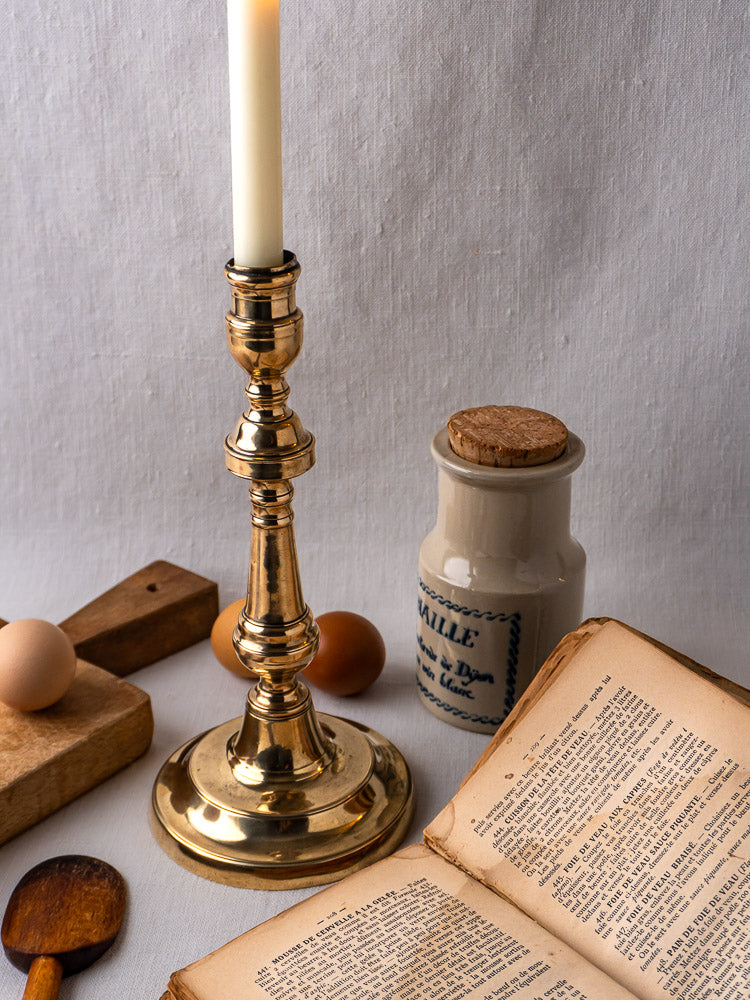 Antique French bronze candlestick with open book, ceramic jar, eggs, and spoon on beige linen.