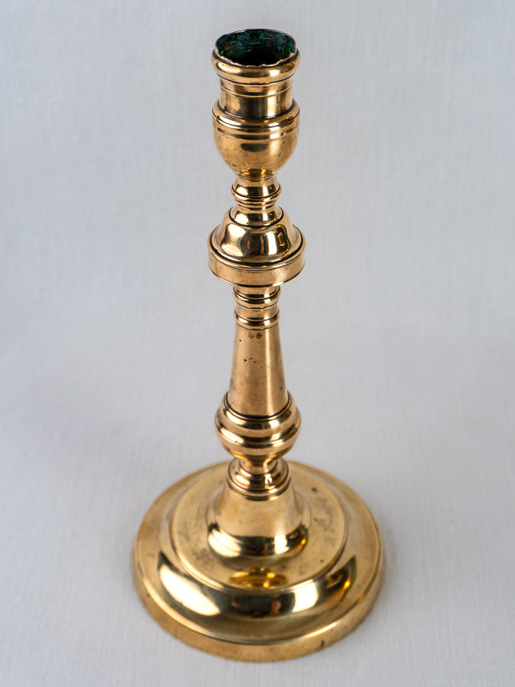 French antique-style polished brass candlestick with turned baluster stem on round base, plain backdrop.
