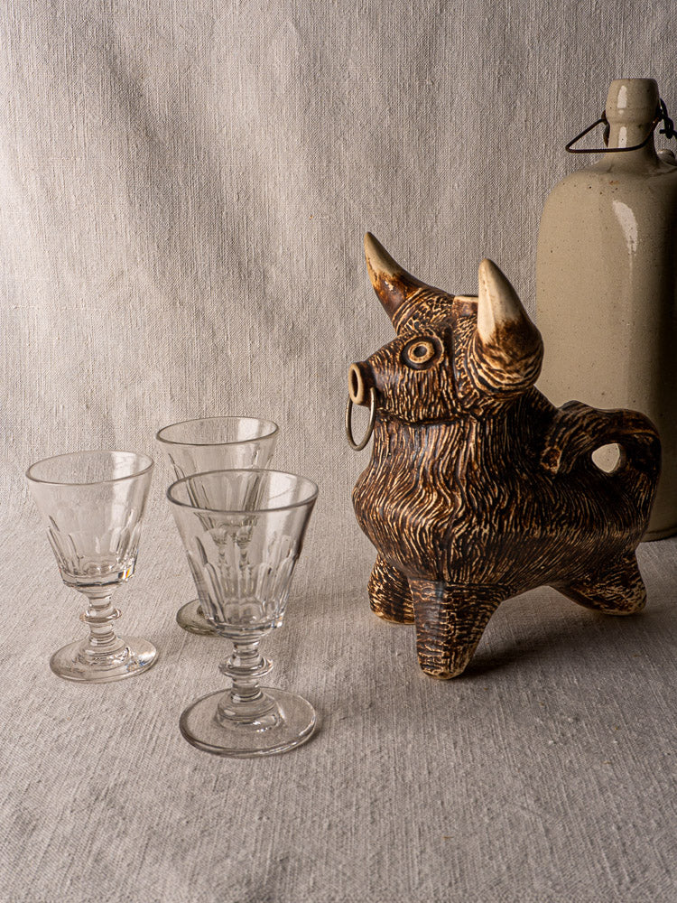 Vintage French bull zoomorphic ceramic pitcher, circa 1950, with three glass goblets and a beige bottle on gray fabric.