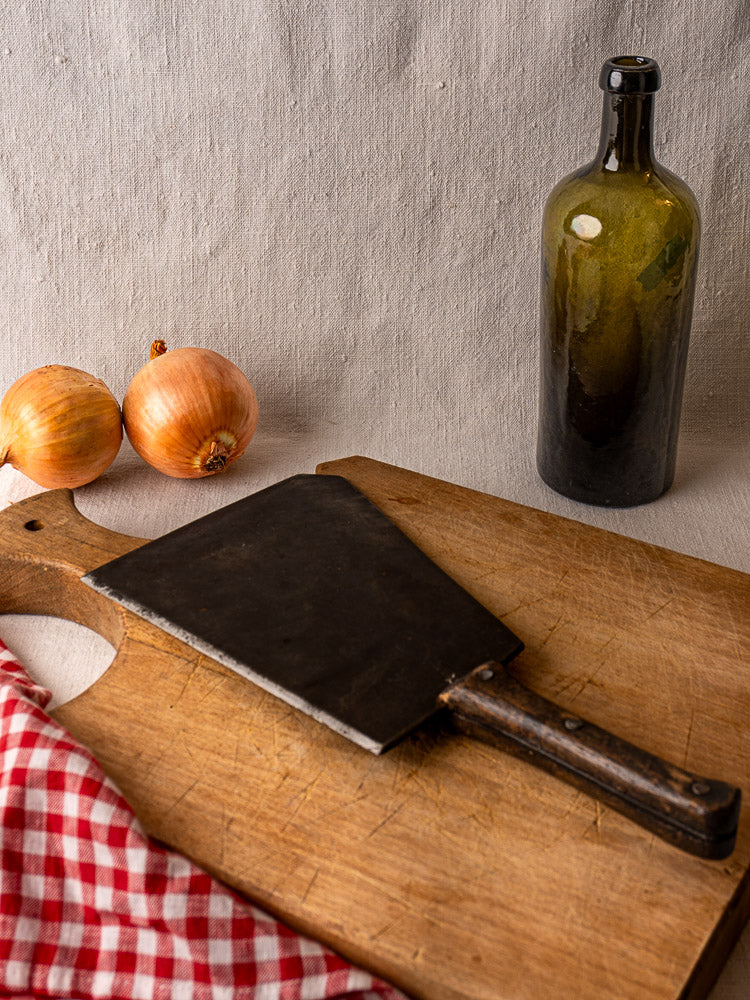 Mid-19th century hand-forged French butcher cleaver on wood cutting board with onions, green bottle, and checkered cloth nearby.