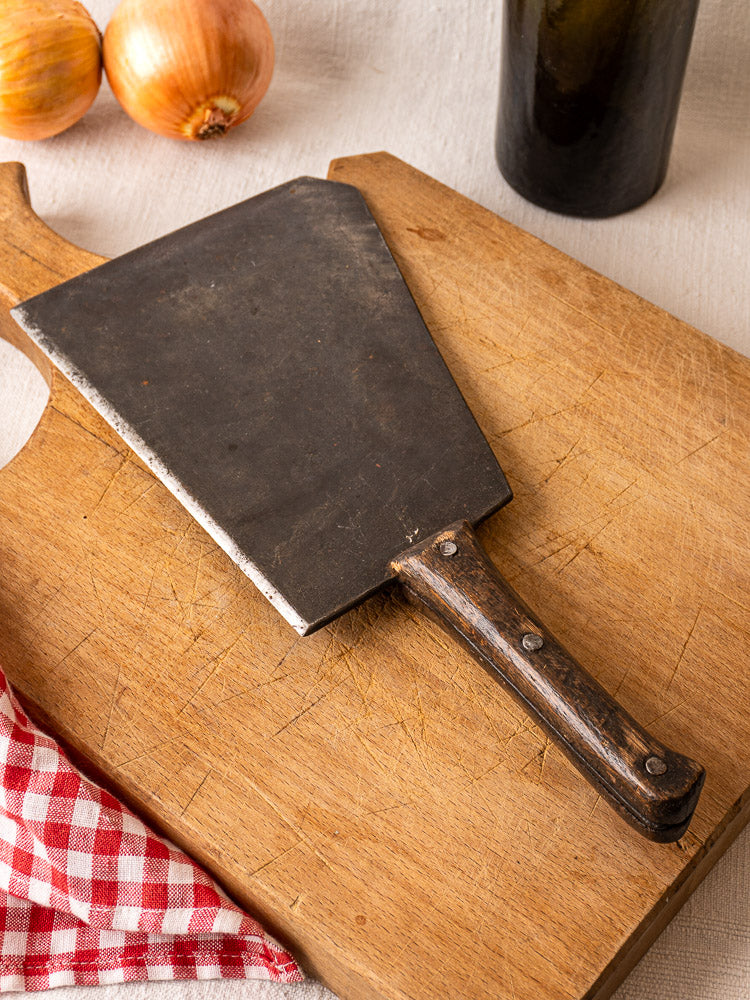 Hand-forged antique French butcher cleaver with wooden handle on cutting board, beside onions and checkered cloth.
