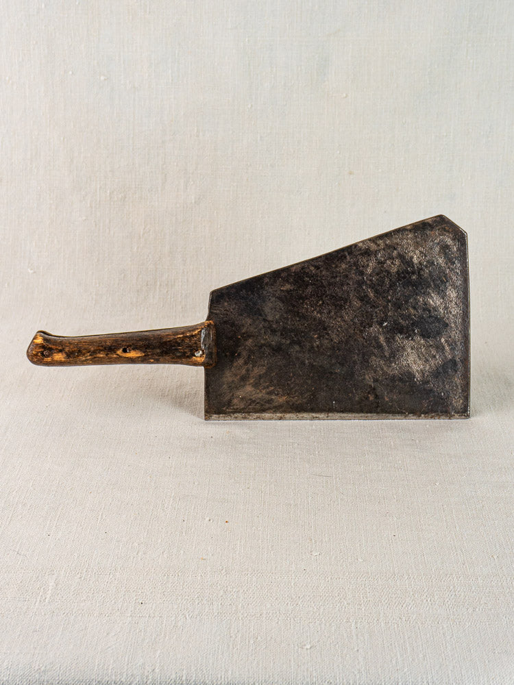 Antique French butcher cleaver with dark weathered blade and wooden handle, shown upright on neutral fabric background.