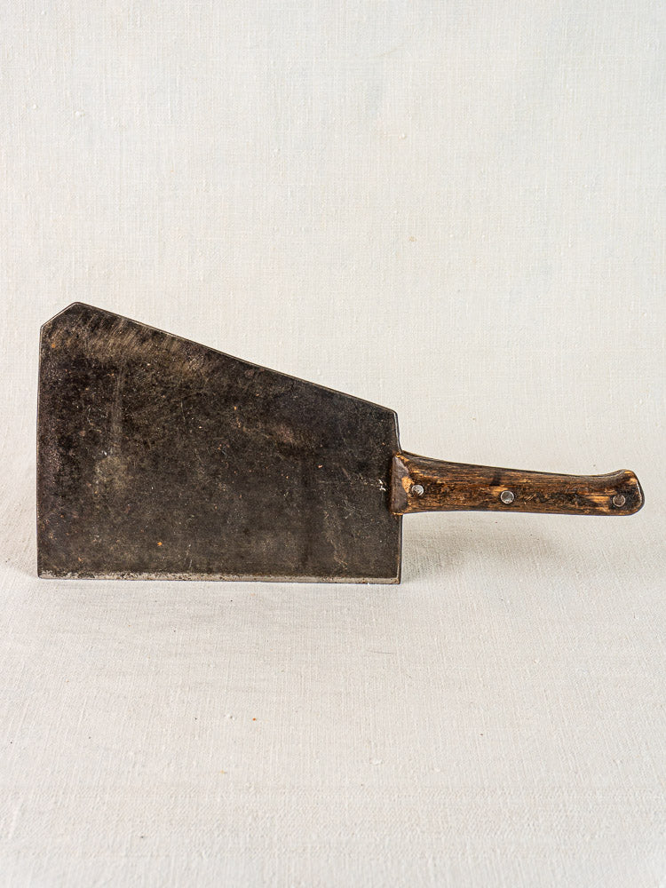 Antique French butcher cleaver with wide dark metal blade and worn wood handle on a light textured surface.