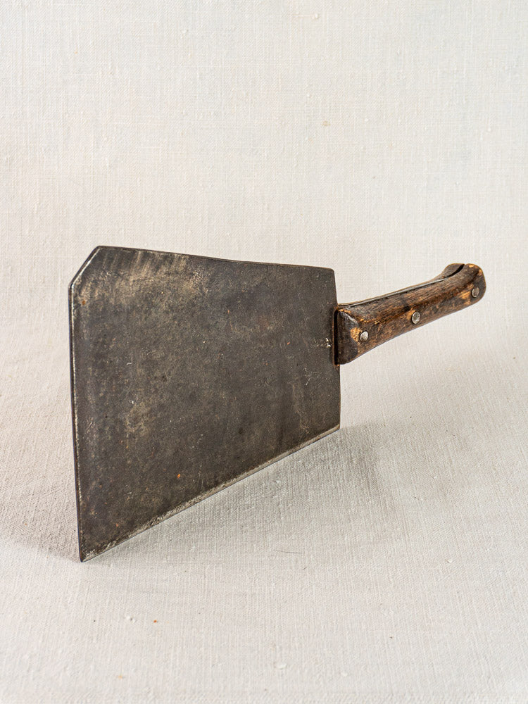 Antique French butcher cleaver with hand-forged, dark blade and wooden handle displayed on a light-textured surface.