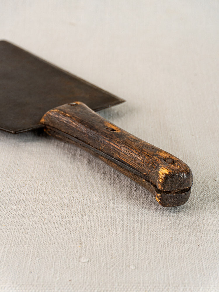 Antique French hand forged butcher cleaver with worn wooden handle and dark blade on light textured fabric surface.