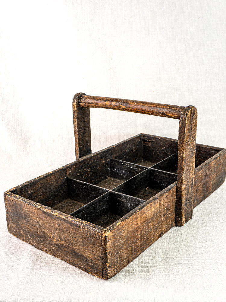 Antique French oak tool tote with hand-forged nails and six compartments on light fabric background.