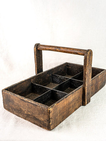 Antique French oak tool tote with hand-forged nails and six compartments on light fabric background.