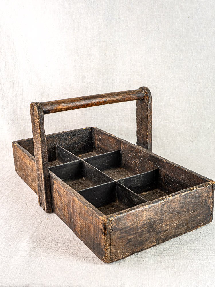 Antique French oak tool tote with hand-forged nails and six compartments on light fabric background.