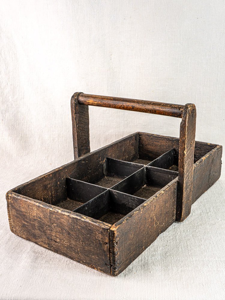 Antique French oak carpenter tote with hand-forged nails, six compartments, and sturdy handle on light textured surface.