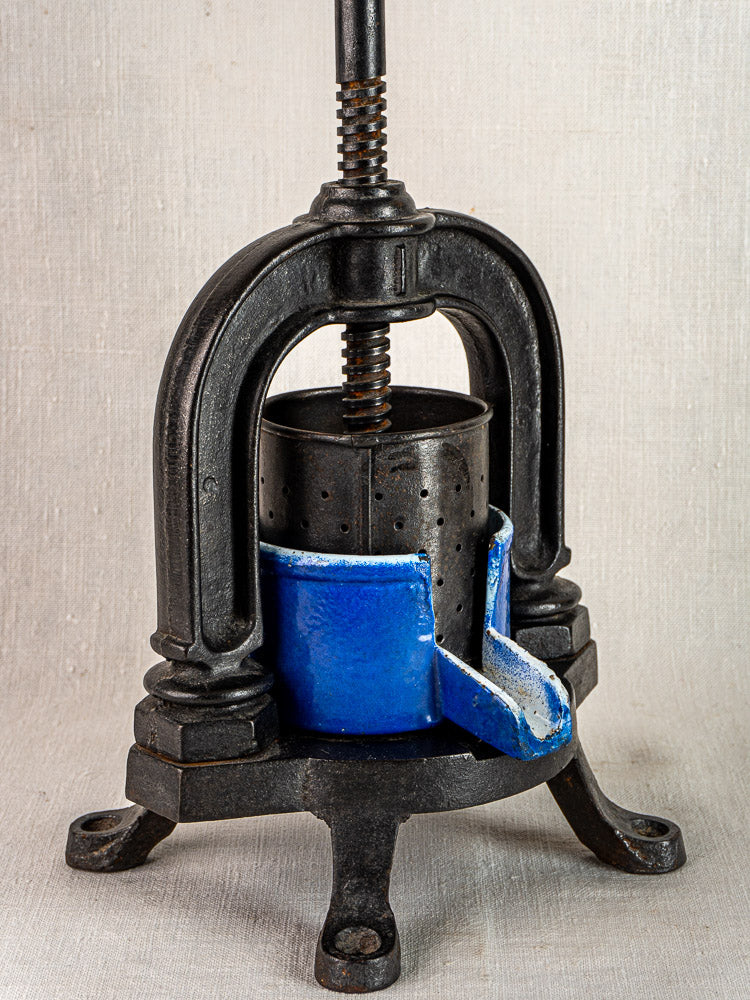 Antique French cast iron tomato press with blue enamel base, vintage black iron on three legs, neutral background.