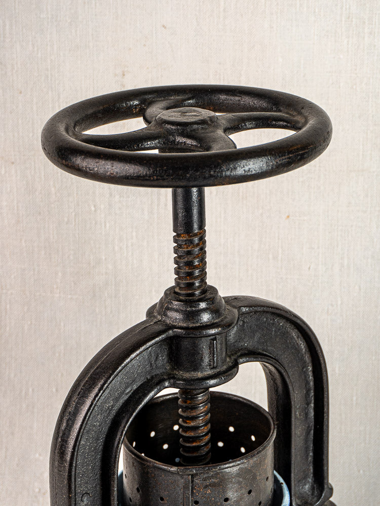 Antique French cast iron tomato press with blue enamel finish, large handle, on neutral fabric background.
