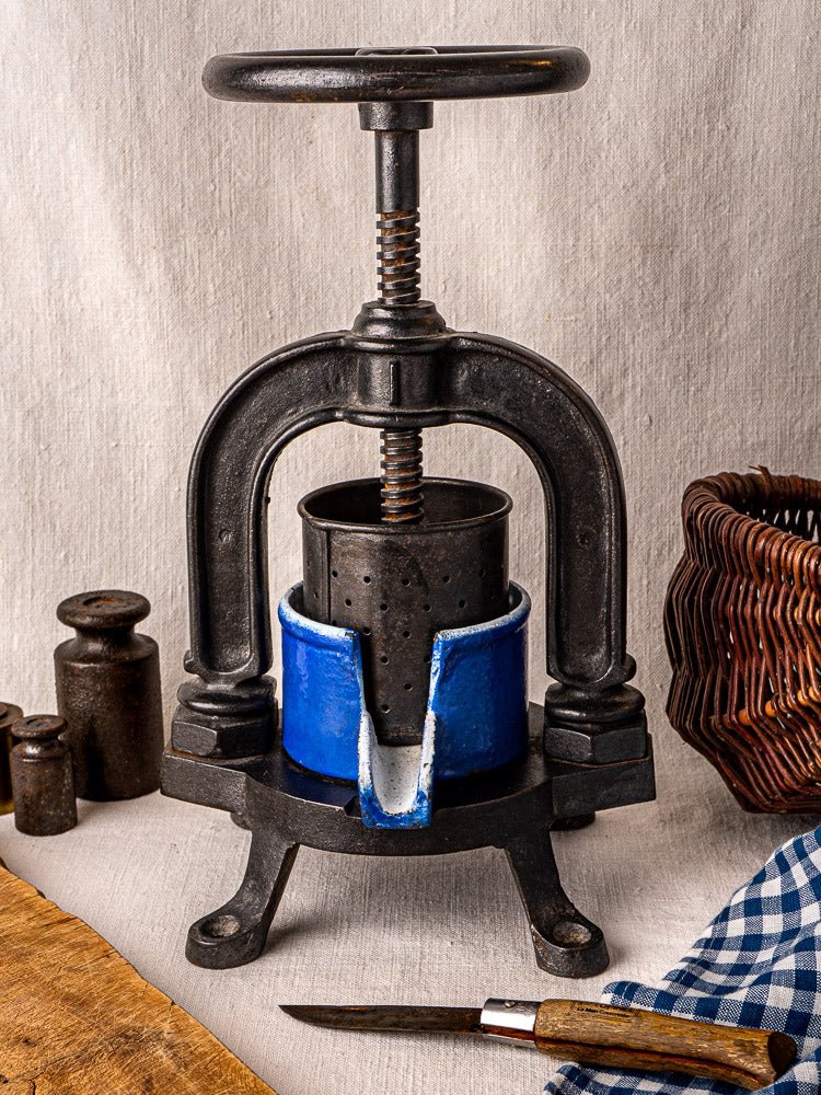 French cast iron tomato press with blue enamel container and black screw handle on neutral background.