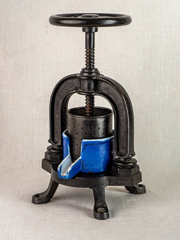 French cast iron tomato press with blue enamel container and black screw handle on neutral background.