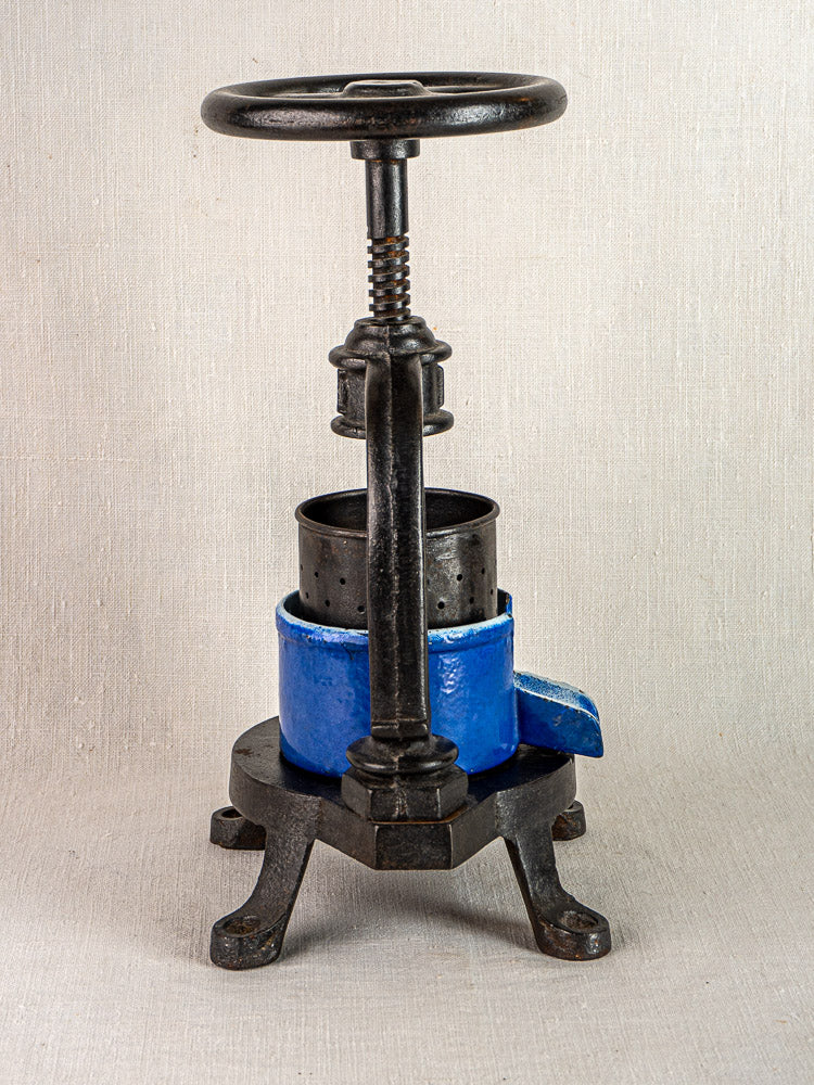 French cast iron tomato press with black metal frame and blue enamel cylinder on neutral surface.