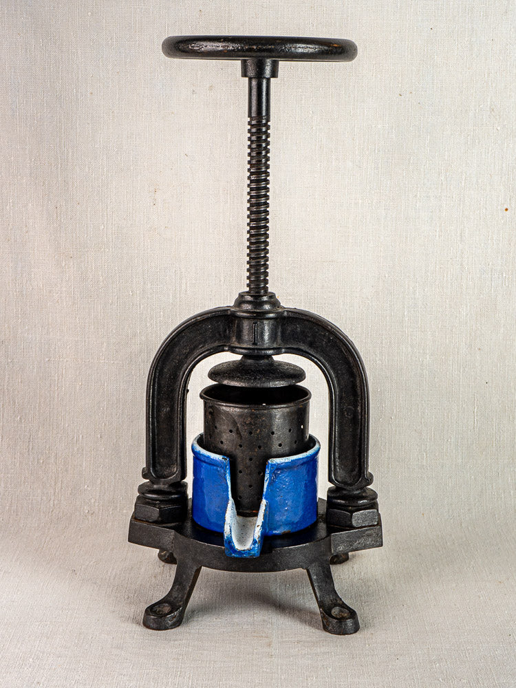 French cast iron tomato press with large screw handle and blue enamel container on neutral background.