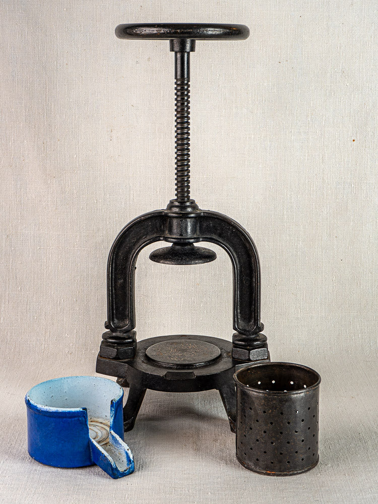 French cast iron tomato press with screw handle and blue enamel container, on fabric backdrop.