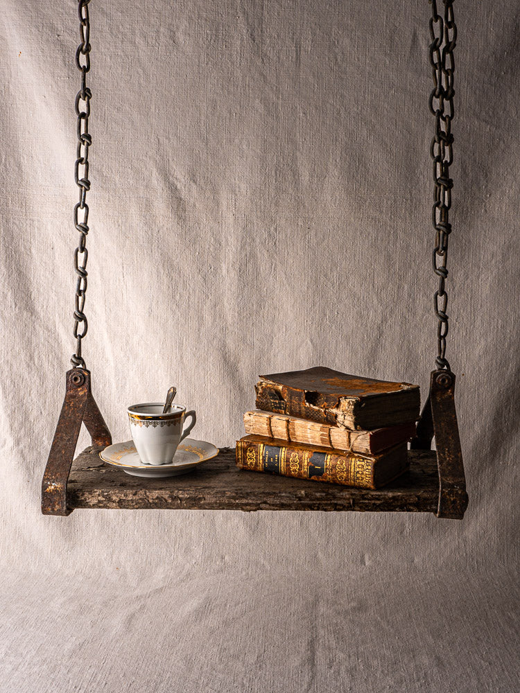 Antique French child’s swing in weathered wood and iron with books and teacup on fabric backdrop.