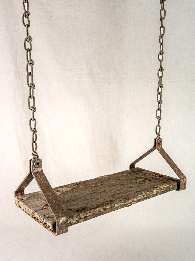 Antique French child’s swing, c. 1900, with weathered wood seat and iron chains on light fabric background.