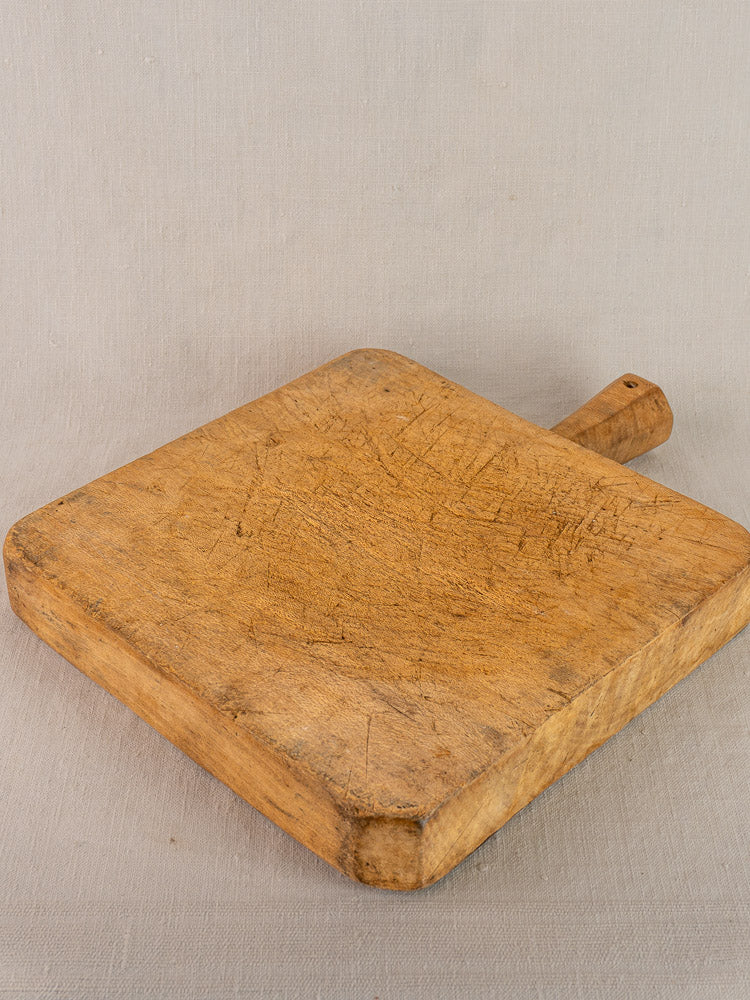 Square antique French beech chopping board with six-sided handle, hand carved and worn, resting on pale fabric.