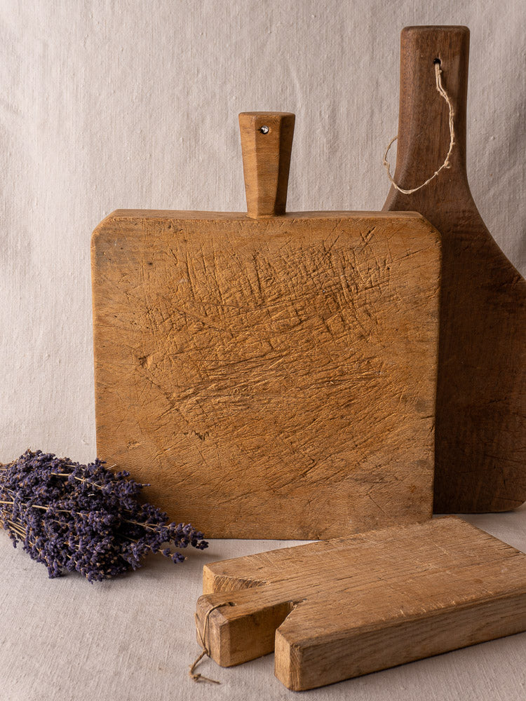 Antique French square chopping board with six-sided handle on beige fabric, dried lavender accent in corner.