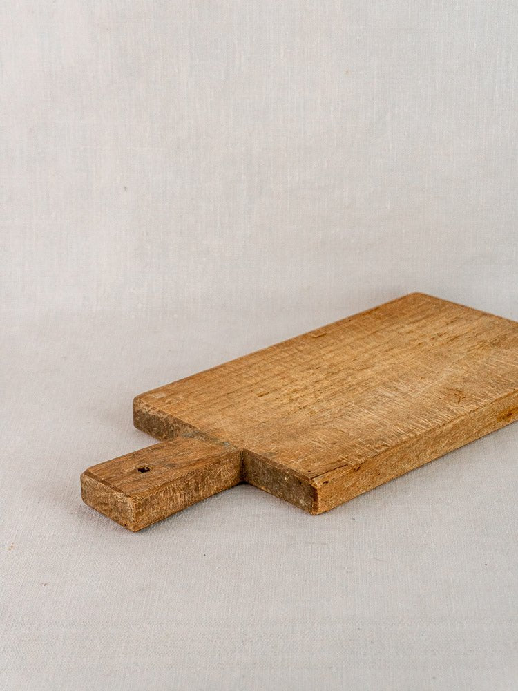 Antique 19th-century French hardwood cutting board with a short handle on beige fabric surface.