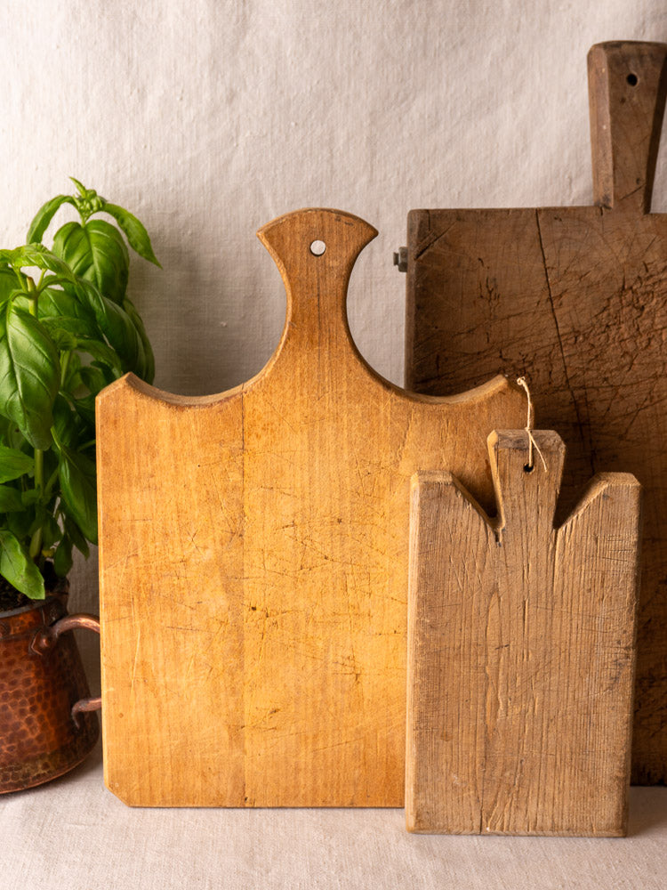 Antique French cutting board with fan handle, rustic boards, and basil in a copper pot against a light backdrop.