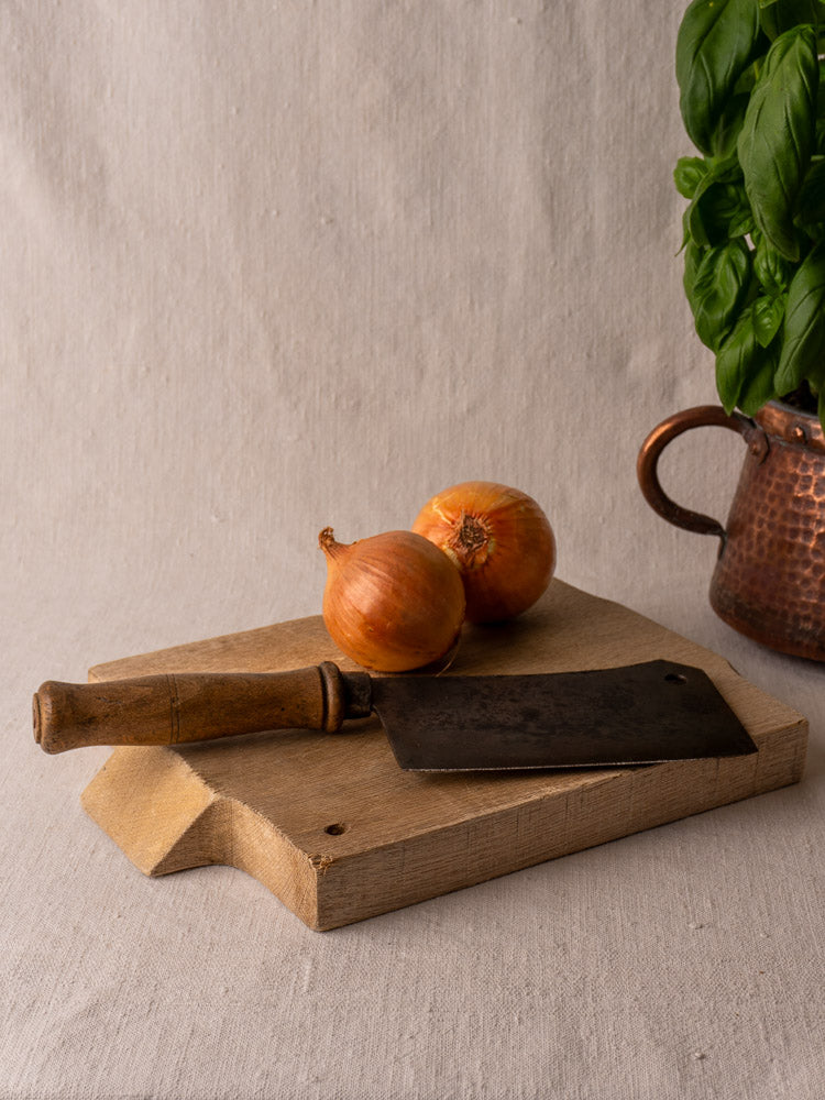 Antique French solid oak cutting board with vintage cleaver, onions, and copper pot of fresh basil on beige cloth.