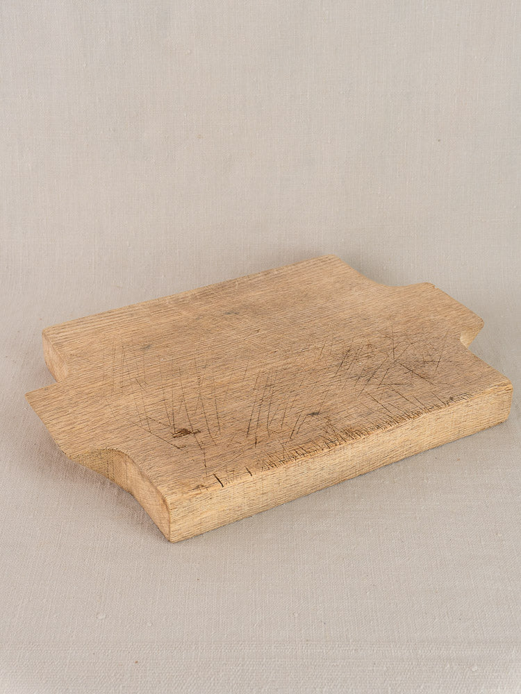 Antique French solid oak cutting board with rounded corners and knife-marked surface on light beige fabric.