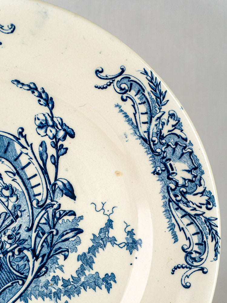 Cream-colored ceramic plate with blue floral and ornate rim patterns, gently worn terre de fer.