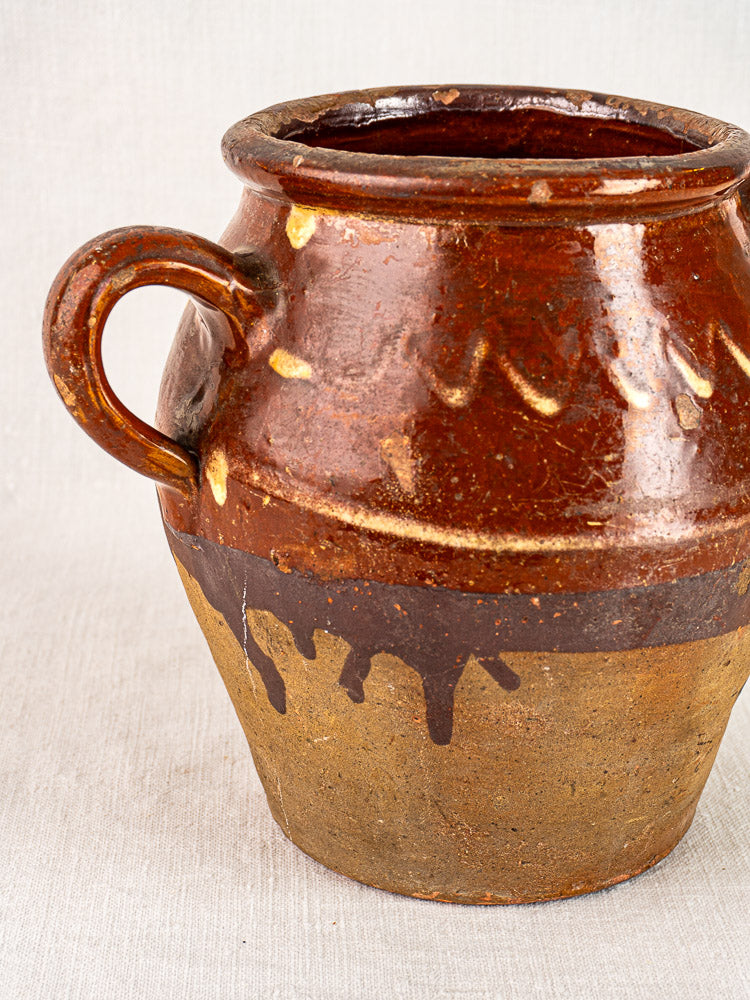 Antique French confit pot with brown glaze, yellow slip rim, single handle, rounded body; on neutral textured surface.