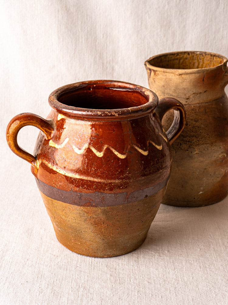Antique French confit pot with brown glaze and yellow slipwork beside a matte ceramic handled pot on light textured surface.