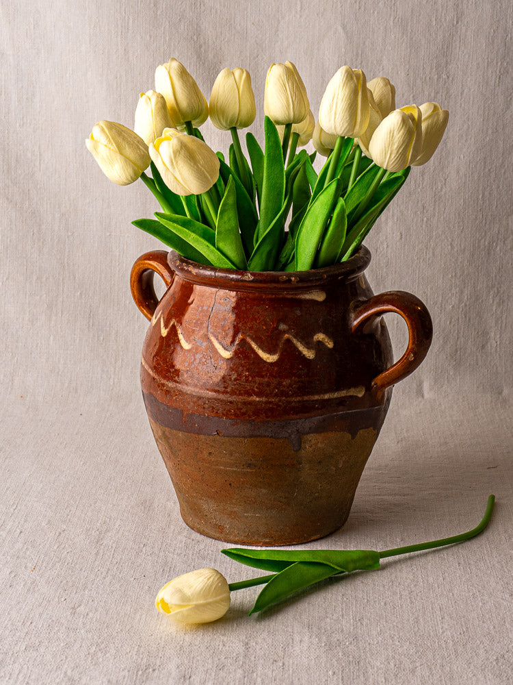 Antique French confit pot with brown glaze and yellow slipwork, filled with white tulips on beige fabric.
