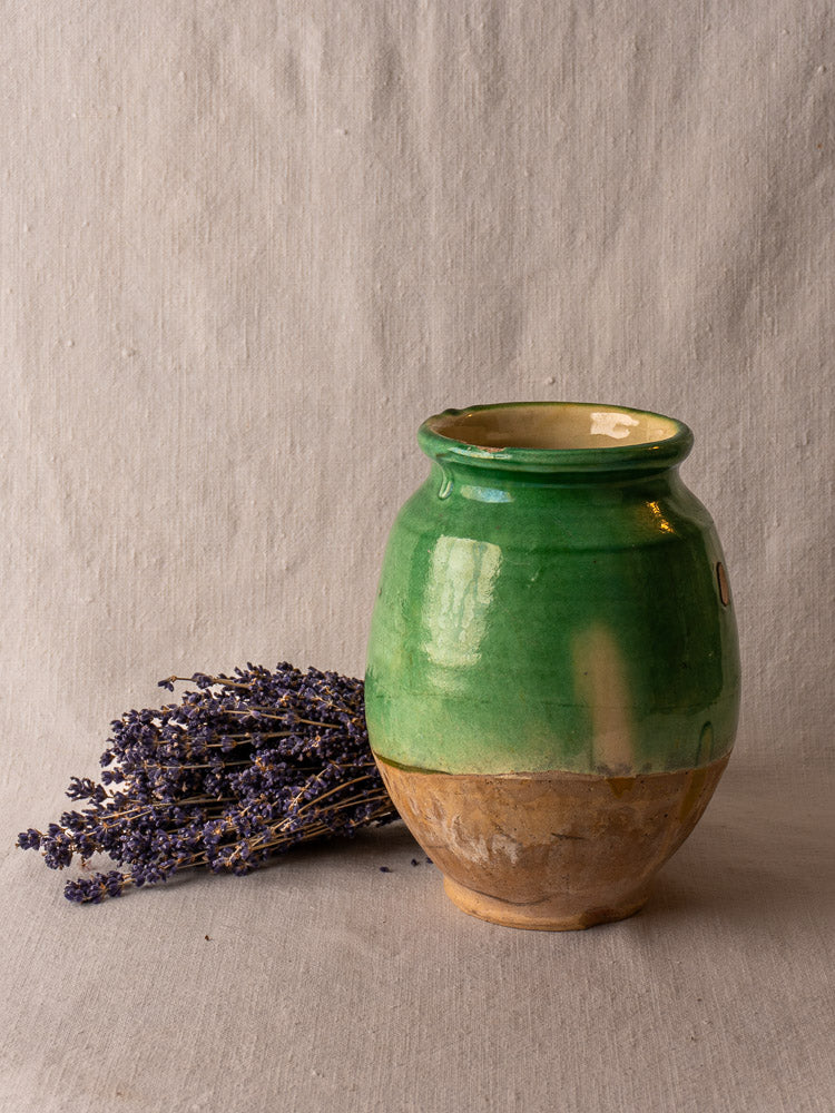 Antique French confit pot with green glazed terracotta finish on beige fabric next to dried lavender.