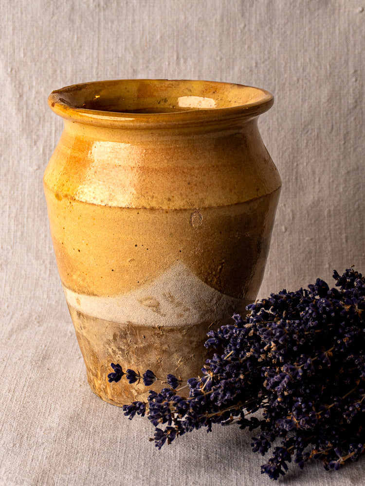 Yellow glazed antique French terracotta confit pot beside dried lavender on beige fabric surface.