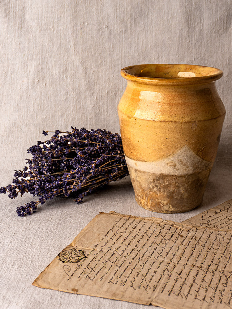 Antique French confit pot with yellow glazed terracotta, displayed with dried lavender and letter on textured fabric.