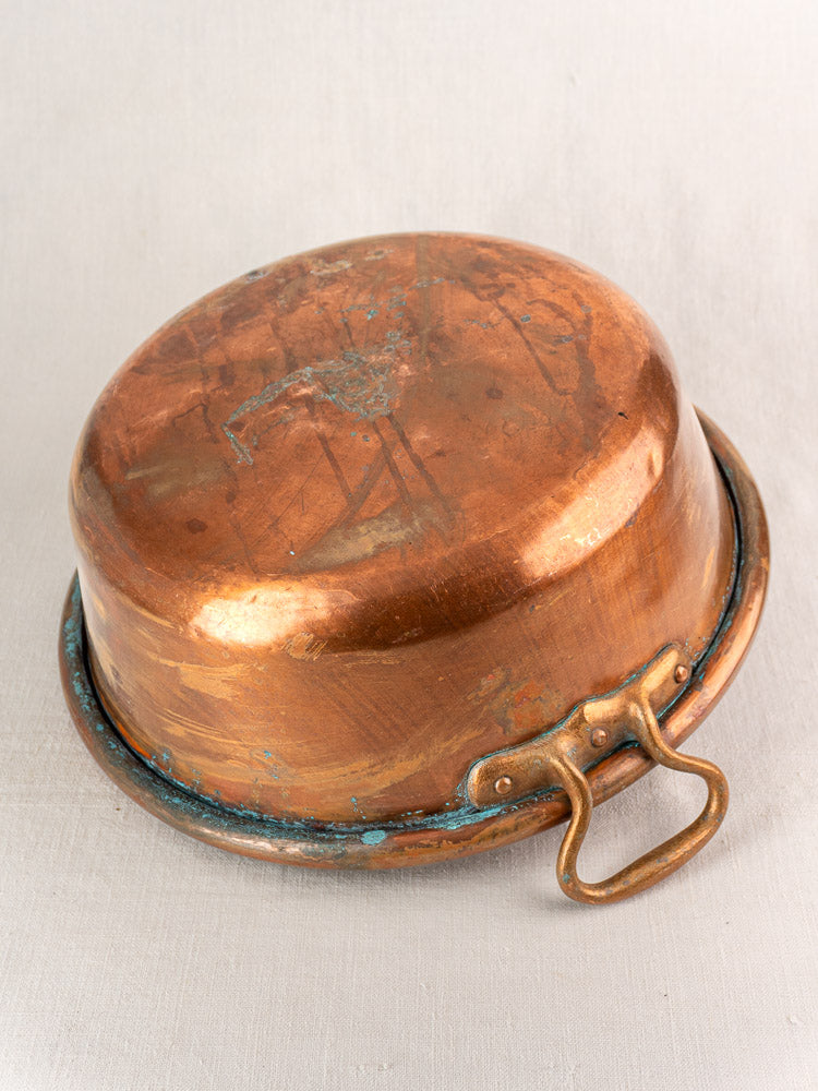 Antique French copper basin with brass handle, tarnished patina, displayed upside down on light fabric.